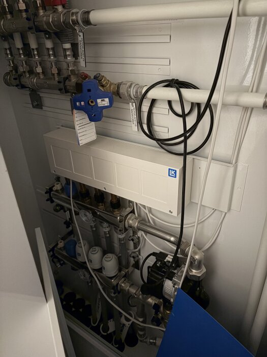 Fördelarskåp för vattenburen golvvärme med ventiler, rör och kopplingar samt en blå komponent och kablar i ett radhus.