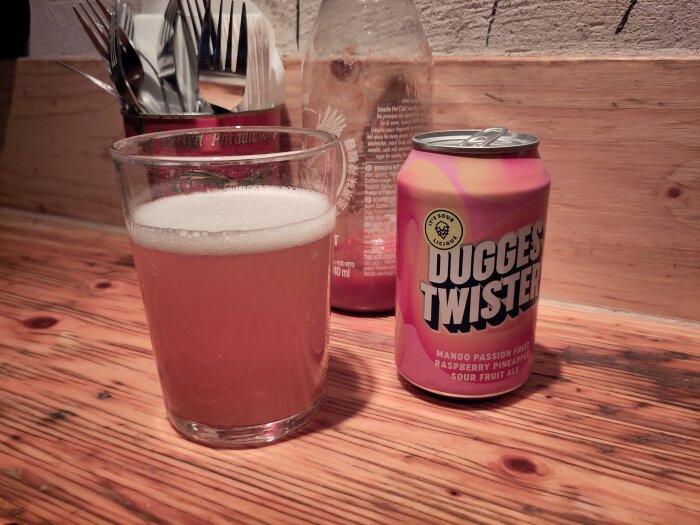 Ett glas med ljus dryck och en burk Dugges Twister Mango Passion Fruit Raspberry Pineapple Sour Fruit Ale på ett bord av trä.