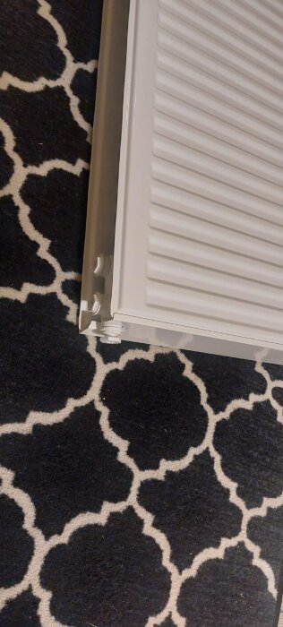 Vattenburet värmeelement monterat mot en mönstrad matta, visar radiatorns anslutningsrör och ribbad design.