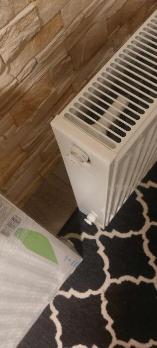 Vit vattenburet värmeelement framför en stenvägg, sedd uppifrån, med termostatventil och rörkoppling.