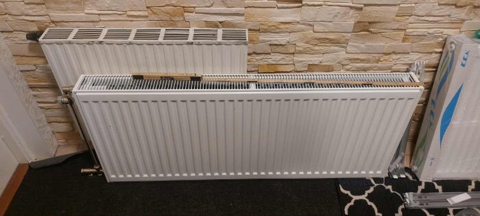 Vattenburet värmeelement lutande mot en tegelvägg, med anslutningsrör synliga på ena sidan.