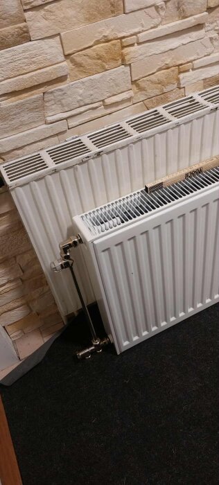 Vattenburet värmeelement lutad mot vägg med rör och ventiler synliga, framför en stenliknande vägg och svart golvmatta.