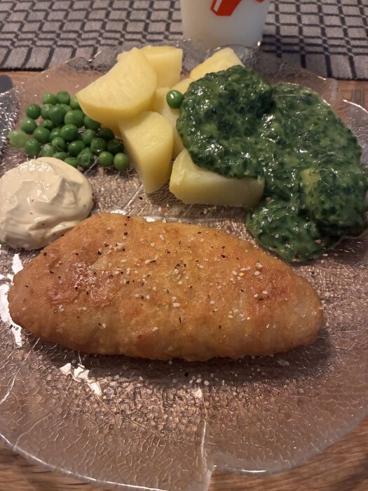Tallrik med panerad fisk, kokta potatisar, ärtor, stuvad spenat och en klick sås.