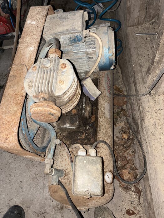 Gammal och rostig luftkompressor med ventiler och kablar synliga, placerad på ett dammigt golv.