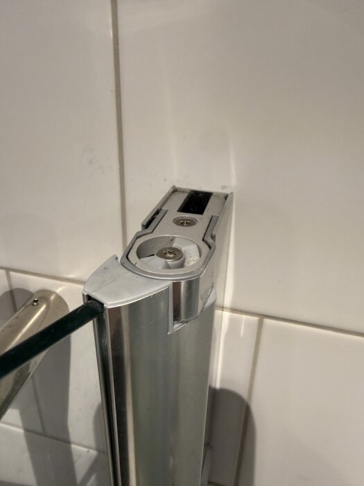 Gångjärn till glasduschdörr på övre del, delvis isärtaget, mot vit kakelvägg.