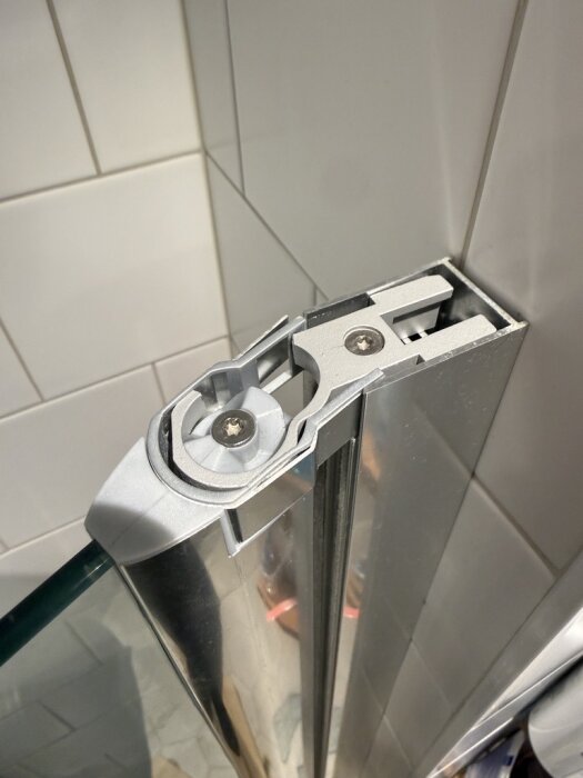 Överdel av duschdörr i glas med intakt gångjärn på en kaklad vägg, fokus på metallfäste och skriven skruv.