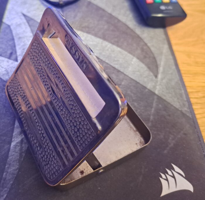 Metallåda för pappersnäsdukar med texten "Nassau", placerad på en musmatta med logga, i bakgrunden en fjärrkontroll.