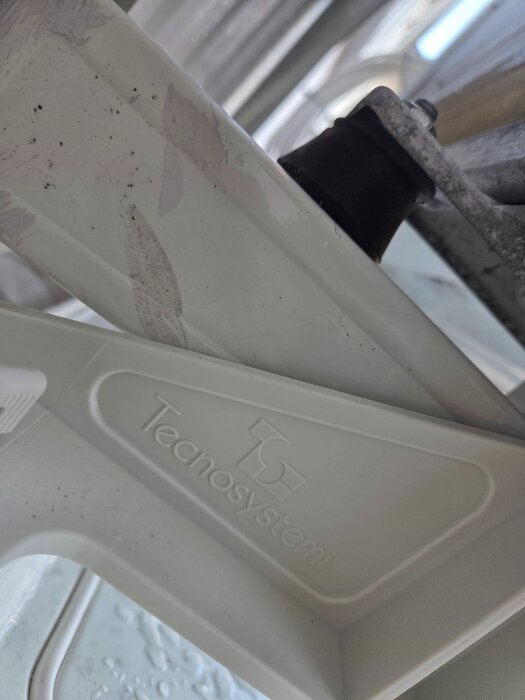 Närbild av en luftvärmepumps stödfot med texten "Tecnosystemi" synlig på en vit plastdel. En svart gummidämpare sitter på en metallkonsol.