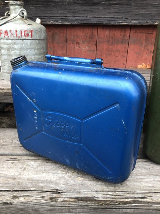 Blå vintage bensindunk märkt "Stopp Nix" på en träyta, med en behållare märkt "Farligt" i bakgrunden.