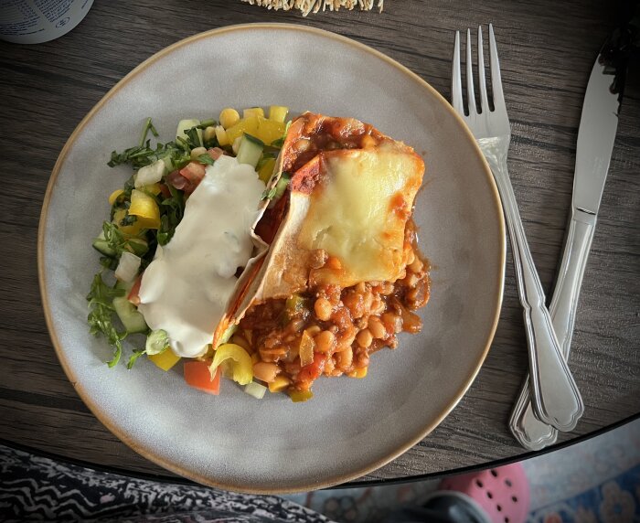 Köttfärs och bönröra i tortilla, gratinerad med ost, serverad med sallad och vit dressing på tallrik.