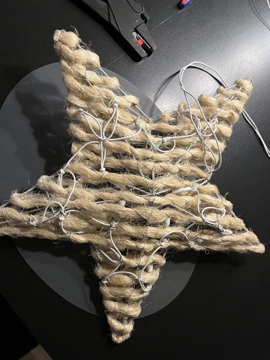 Flätad stjärna av rep dekorerad med vita LED-slingor, fotograferad ovanifrån på ett svart underlag.