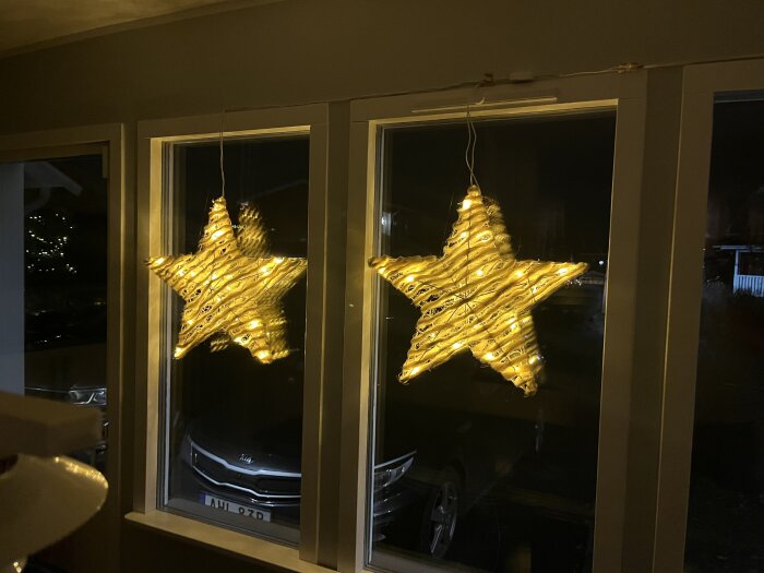 Två stjärnformade ljusinstallationer med LED-lampor hänger framför ett fönster, lysande med ett varmt gult sken i mörkret.