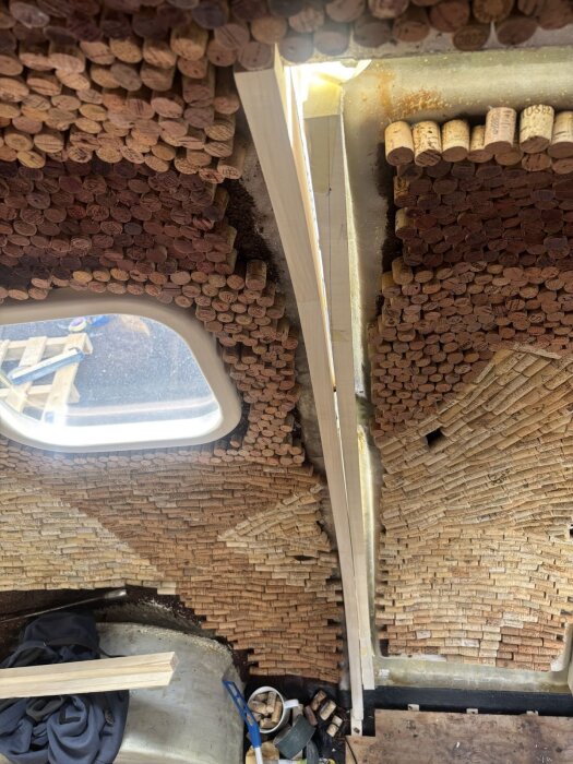 Vägg täckt med vinkorkar och träram under arbete. Korkar arrangerade på vägg, synlig ljuskälla ovan, träbit centralt och byggverktyg på golvet.