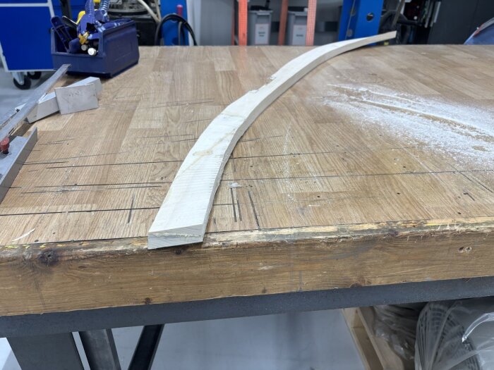 En böjd träbit med twist ligger på en arbetsbänk i en verkstadsmiljö, omgiven av verktyg och sågspån.