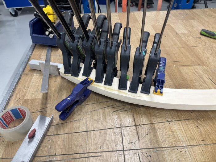 Träbit pressad med flera tvingar på ett arbetsbord i en verkstadsmiljö, omgiven av verktyg och tejp.