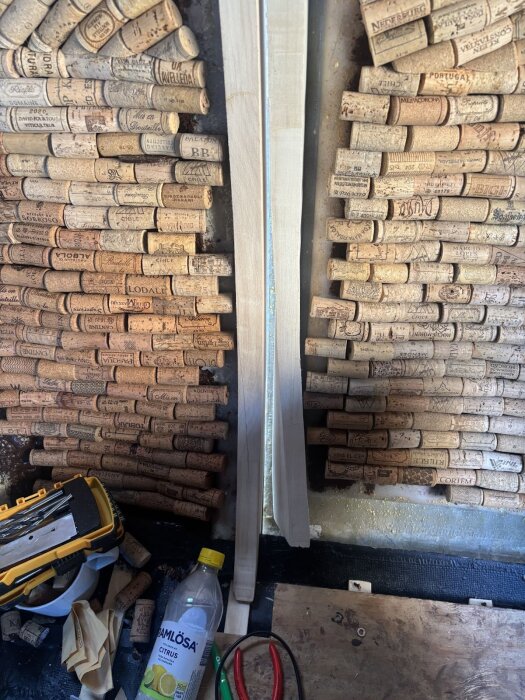 Trävägg med vinkorkar, två träbitar i mitten, verktygslåda och flaska på golvet.