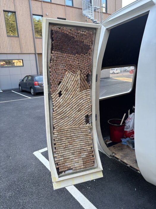 Öppen dörr med konstruktion av vinkorkar och trä, monterad på en vit släpvagn, med byggnad och parkerade bilar i bakgrunden.