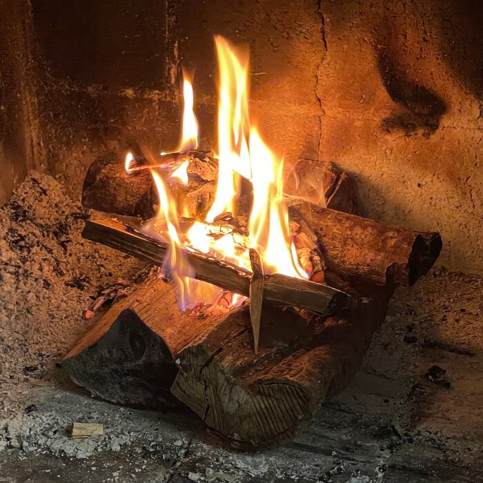 Öppen eld i eldstad, med grova vedträn i botten och mindre stickor överst; lågor som brinner klart och varmt.