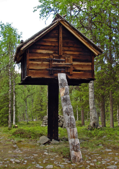 En traditionell njalla byggd på en hög trästam i en skog, med stocktak och trästege framför.