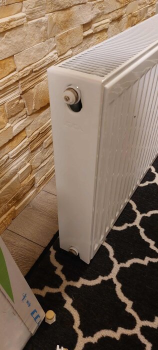 Radiator med anslutningar på vardera sida, står mot en vägg med stenimitation och en mönstrad svartvit matta på golvet.