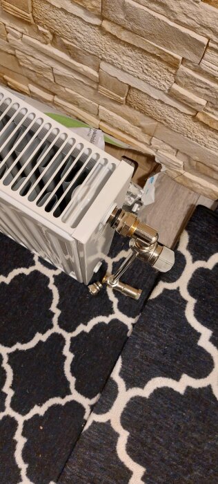 Värmeelement mot en vägg med stenimitation och anslutet rör arbete. Matta med vitmönstrad design syns under.