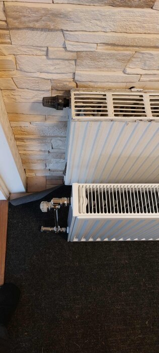 Radiator lutad mot en vägg av stenplattor, visar ventil och anslutningsrör.