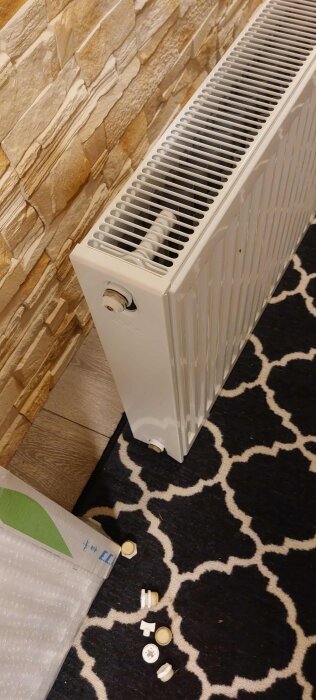 Vit radiator vid stenvägg på mönstrad matta, olika radiatorbeslag synliga på golvet.