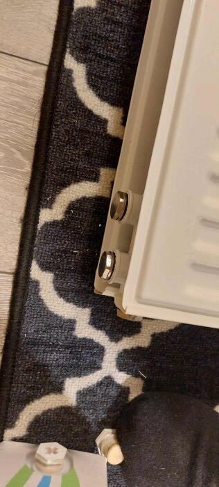 Närbild av en radiator och dess anslutningar, delvis på en matta med geometriskt mönster.