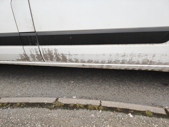 Smutsig bilsida efter misslyckad tvätt, tydliga smutsfläckar längs den vita karossen nära trottoarkanten.
