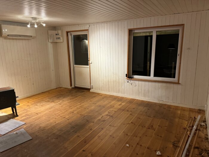 Tomt rum med vitmålad träpanel, trägolv, fönster och altandörr. Syns luftkonditioneringsenhet och spotlights i taket.