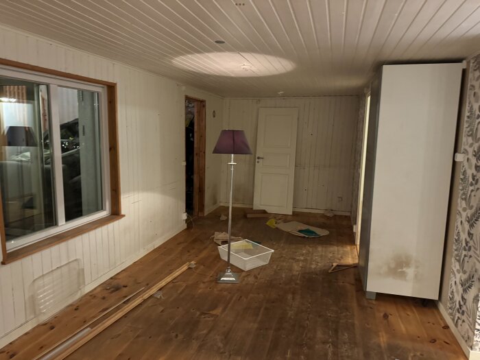 Ett rum under renovering med trägolv, vita väggar och takpanel. Ett fönster till vänster, en golvlampa och några spridda föremål ligger på golvet.