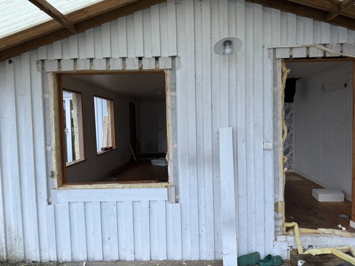 Vitt trähus under renovering med två stora hål för fönster eller dörrar, isoleringsmaterial syns runt kanterna.