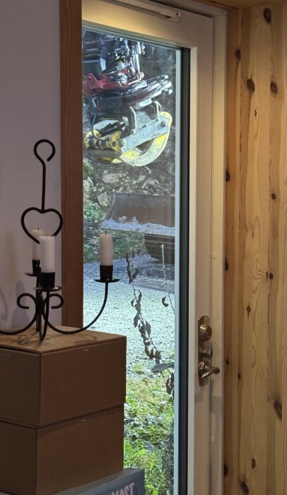 Grävmaskin syns genom glasdörr tillsammans med flyttlådor och ljusstake inomhus.