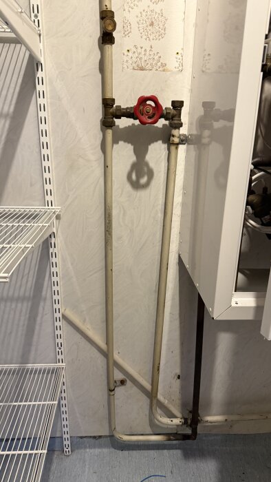Rörinstallationer i en tvättstuga med ventiler och rör mot en ljus vägg, hylla till vänster och del av skåp till höger.
