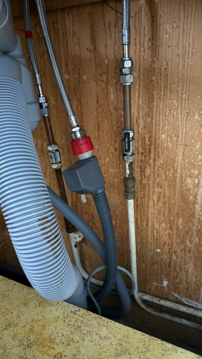 Rörinstallationer i tvättstuga med kopplingar och flexibla slangar mot en trävägg.