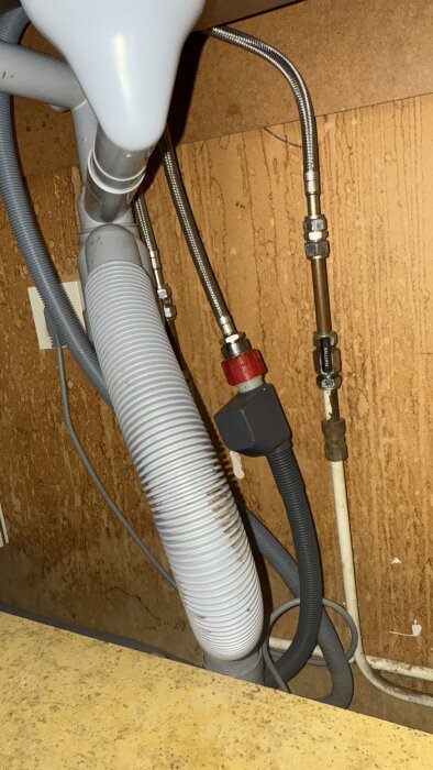 Rör och slangar under diskbänk i tvättstuga, inklusive en vit plastslang och ett grått avloppsrör, visas mot en träpanelbakgrund.