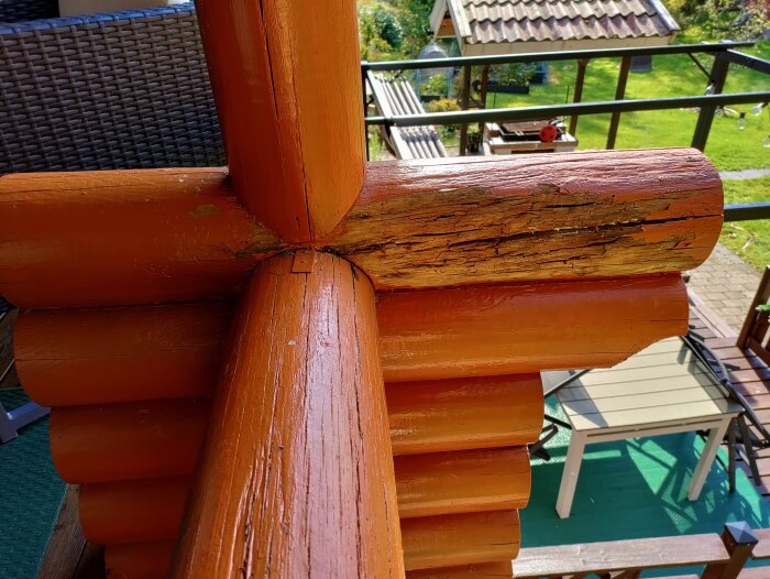 Rötskadat timmerräcke med synliga sprickor i ett timmerhus, del av en byggnadsdetalj, omgiven av utemöbler och grönska.