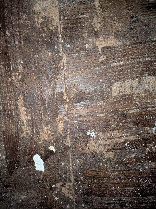 Närbild av en masonitskiva med limrester och sliten yta, efter att en gammal plastmatta tagits bort från golv under renovering i ett äldre hus.