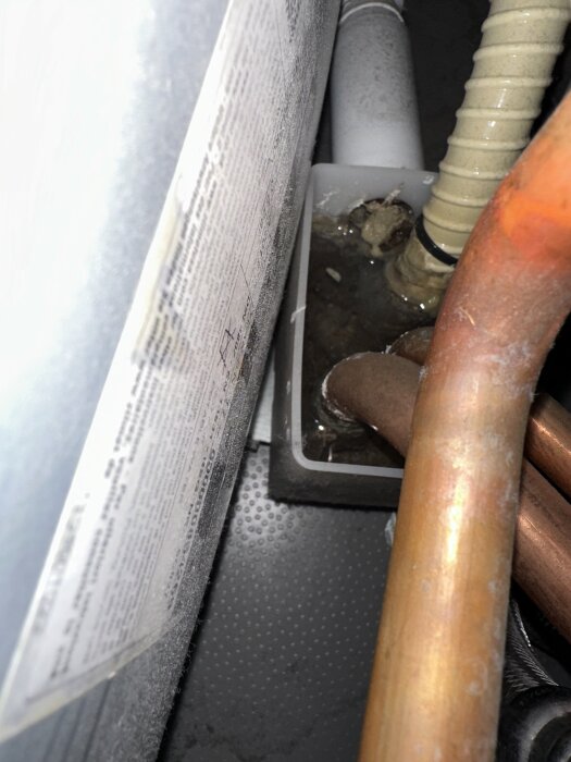 Vit-gul slang med koppar- och plastledningar i en koppling, möjligen igensatt, med smutsigt vatten.