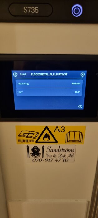 Display på värmepump med inställningar för flödesinställning och DUT på -20 grader, samt en varningsetikett och företagslogotyp.