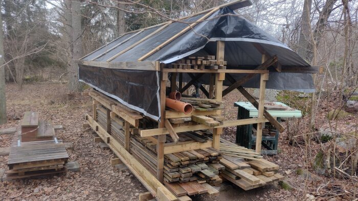 En träkonstruktion förvarar byggmaterial under en presenning i en skogsmiljö.