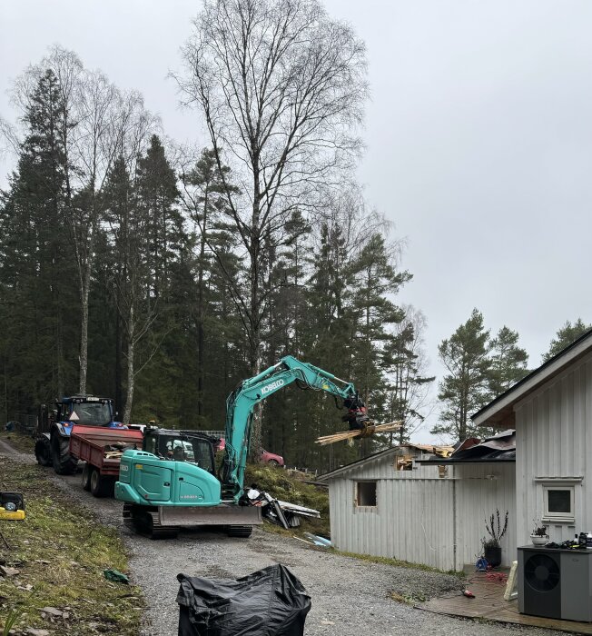 Grävmaskin river en liten vit stuga bredvid en skog, med en traktor och släp som står parkerade i bakgrunden.