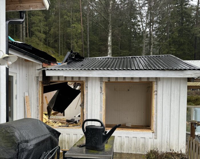 Vit stuga under rivning, med skadat tak och öppet fönster. En svart vattenkanna på ett bord i förgrunden och skog i bakgrunden.