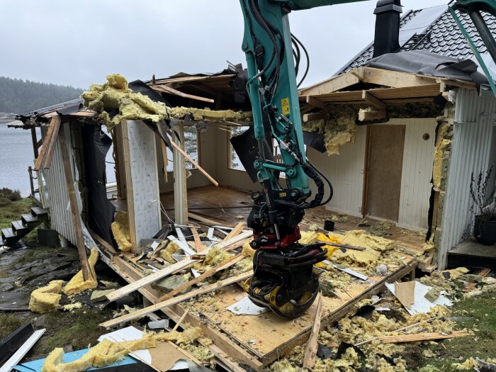 En grävmaskin demolering en gammal stuga vid en sjö, trä och isoleringsmaterial ligger utspritt runt området.