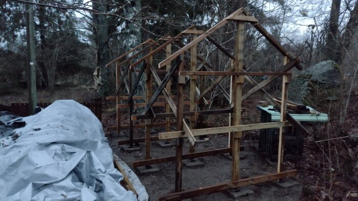 Träram av ett byggprojekt med en presenning i förgrunden, omgiven av skog.