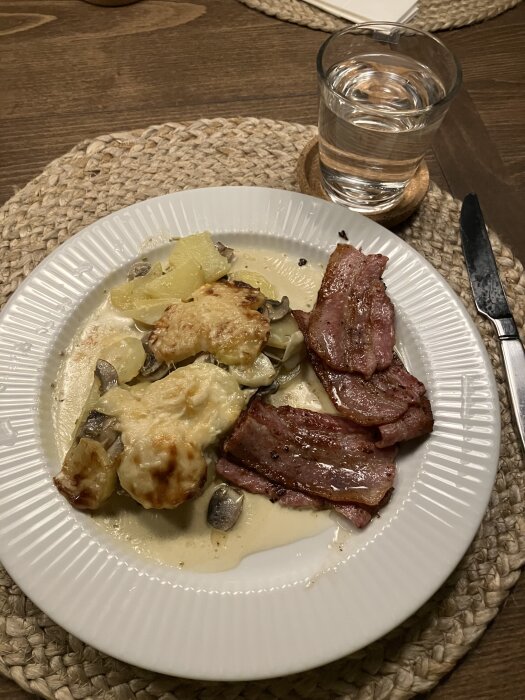 Italiensk potatisgratäng med champinjoner och parmesan, serverad med stekt fläsk, på en vit tallrik. Glas med vatten bredvid.