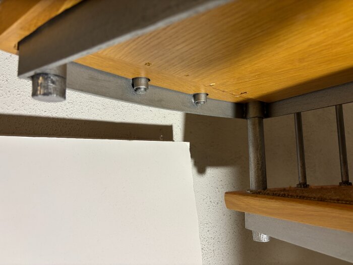 Undersida av spiraltrappa med trästeg och metallkonstruktion, synliga bultar och skruvgängor i närbild, fokus på möjlig reparation.