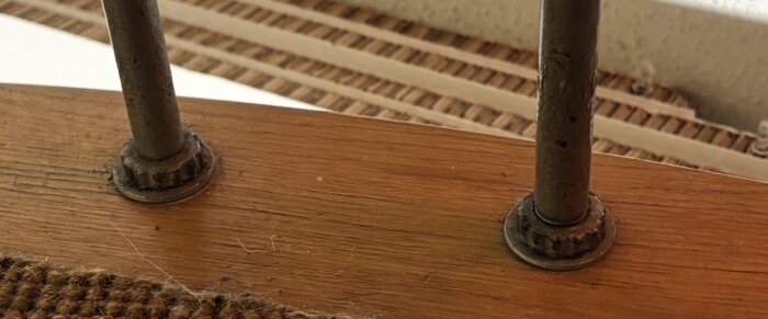 Närbild av två metallstänger fastsatta i en träyta av ett trappsteg, del av en spiraltrappa, med synliga skruvar och mattkant.