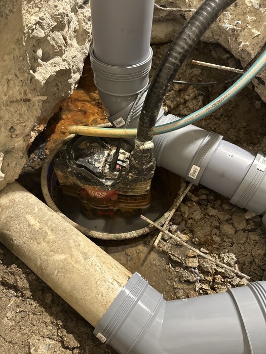 Provisorisk pumpbrunn i källare med grå avloppsrör och synlig vattenansamling under betonggolv.