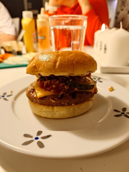 Hamburgare med ost, lök, saltgurka och dressing på en tallrik. I bakgrunden syns senapsflaska och glas med vatten.